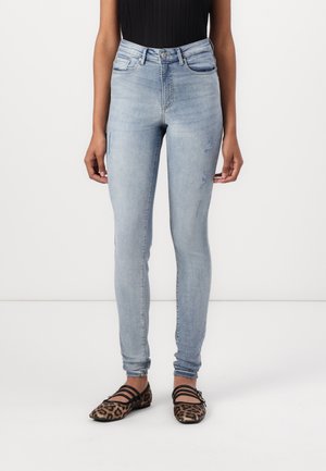Jeans skinnies de tiro alto en azul claro con un ligero desgaste en la parte superior de los muslos. Llevados con unas chanclas negras con tiras que presentan un patrón de leopardo.