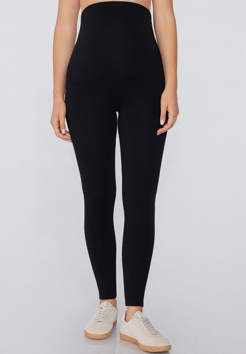 Leggings noirs taille haute en tissu extensible. Présentent une texture lisse et une coupe ajustée, assortis à des baskets de couleur claire.
