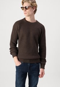 Donkerbruine gebreide trui met een ribbelstructuur, ronde halslijn en raglanmouwen, gecombineerd met donkerblauwe spijkerbroeken. Casual pasvorm.
