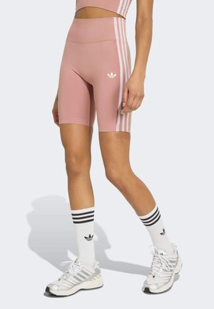 Femme portant un short de cycliste Adidas rose taille haute avec des bandes blanches, des chaussettes blanches Adidas et des baskets blanches, debout devant un fond clair.