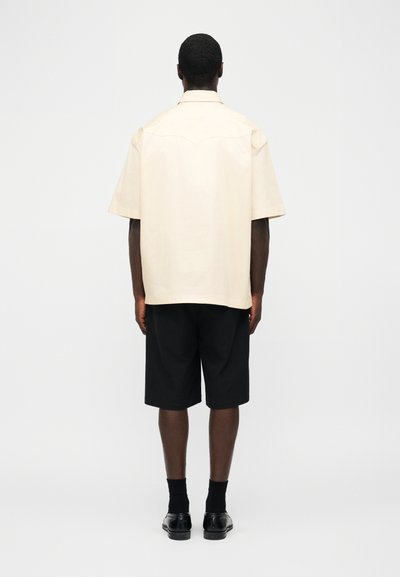 Homme debout de dos, portant une chemise beige à manches courtes, un short noir jusqu'aux genoux, des chaussettes noires et des chaussures habillées noires.