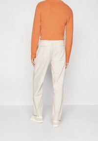 Orange tekstureret sweater, lysebeige bukser med lommer og afslappede hvide sneakers. Neutral baggrund. Fokus på tøjdesign og materiale.