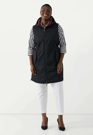 Gilet imbottito navy con cappuccio, caratterizzato da una chiusura con zip e tasche laterali, indossato sopra una camicia a righe e abbinato a pantaloni bianchi e tacchi blu.