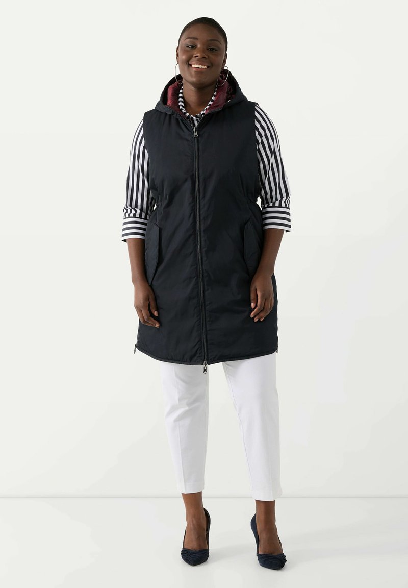 Gilet imbottito navy con cappuccio, caratterizzato da una chiusura con zip e tasche laterali, indossato sopra una camicia a righe e abbinato a pantaloni bianchi e tacchi blu.