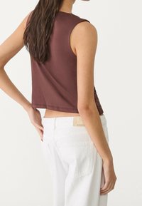 Brązowy dopasowany bezrękawnik z okrągłym dekoltem, zestawiony z białymi dżinsami o wysokim stanie z metką na tylnej części pasa.