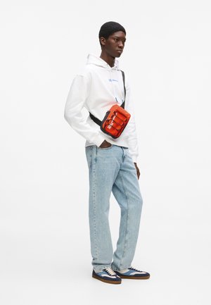 Νέος άνδρας που φοράει λευκό φούτερ με κουκούλα, ανοιχτόχρωμο τζιν, μπλε αθλητικά παπούτσια και πορτοκαλί τσαντάκι χιαστί σε λευκό φόντο.