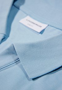 Ljusblå pikétröja med kort krage, tillverkad av texturerad bomullstyg. Lappen är synlig och visar varumärke och storlek.