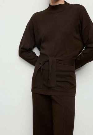 Personne portant un haut en maille à manches longues marron foncé avec une ceinture nouée à la taille et un pantalon large assorti, sur un fond clair uni.