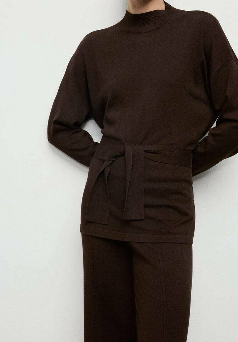 Personne portant un haut en maille à manches longues marron foncé avec une ceinture nouée à la taille et un pantalon large assorti, sur un fond clair uni.