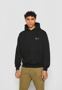 Hoodie en coton noir avec une poche frontale, des poignets et une taille côtelés. Il présente un petit logo blanc sur la poitrine et une capuche avec cordon de serrage.