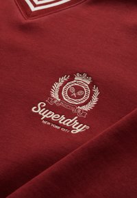 Sudadera burdeos con escote en V, cuello blanco a rayas y logotipo bordado con un diseño de corona y laureles en color crema.
