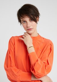 Helles orange Bluse mit langen, gepufften Ärmeln und Knopfdetails an der Schulter. Das Model trägt zudem ein goldenes Armband mit schwarzen Akzenten.