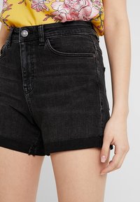 Svarta denimshorts med fransar, med fem fickor och knappstängning, stylade med en blommig gul topp.