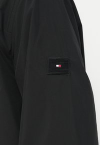 Czarny rękaw kurtki z prostokątną łatą logo Tommy Hilfiger, w kolorach czerwonym, białym i niebieskim, umieszczoną na górnej części ramienia.