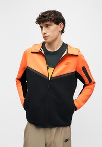 Νεαρός άνδρας με σγουρά μαλλιά που φοράει πορτοκαλί και μαύρο μπουφάν Nike με φερμουάρ, μαύρο μπλουζάκι και μπεζ παντελόνι Nike, με τα χέρια στις τσέπες του μπουφάν.