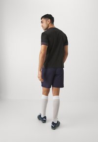 Camisola atlética preta, de mangas curtas com detalhes cinzas, calções azuis-marinho, meias brancas até ao joelho e chuteiras de futebol pretas com detalhes verdes.