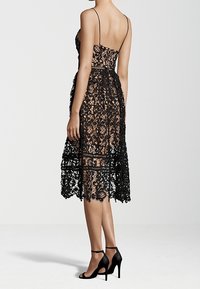 Robe en dentelle noire à bretelles spaghetti, présentant des motifs élaborés, un corsage ajusté et une jupe longue et fluide. Associée à des talons noirs.