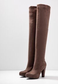 Botas de ante marrón hasta la rodilla con una textura suave, tacón ancho alto y diseño de puntera redondeada. Los detalles de las costuras verticales añaden interés visual.