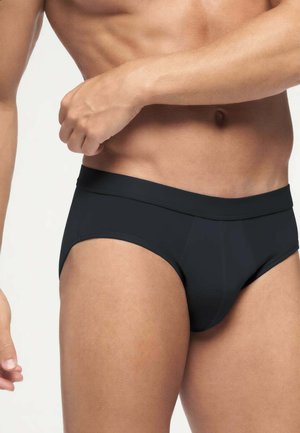 Uomo in forma che indossa slip neri con fascia in vita a media altezza e tessuto con finitura liscia, in piedi su uno sfondo chiaro e uniforme.