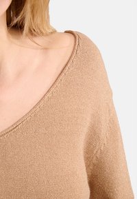 Donna con capelli biondi che indossa un maglione lavorato a maglia beige con un ampio scollo a V che mostra l'area delle clavicole e delle spalle.