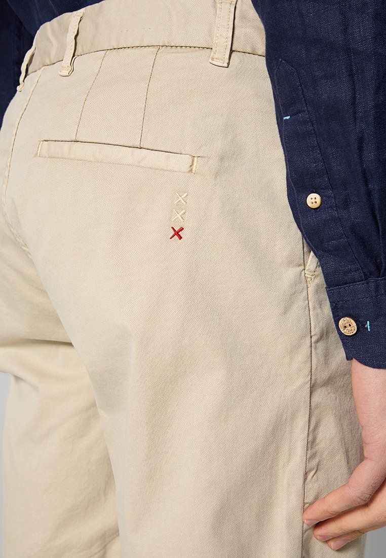 Beżowe spodnie bawełniane mają tylną kieszeń z dekoracyjnym haftem w białe i czerwone kształty X. Materiał ma gładką fakturę.
