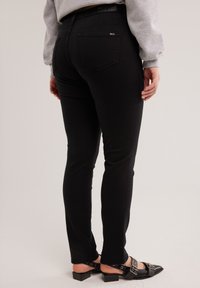 Egy személy fekete, szűk szabású farmert és fekete hegyes orrú cipőket, csattokkal visel, bézs háttér előtt áll.