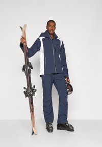 Kék és fehér kabátos síöltözet cipzáras záródással, kék nadrággal párosítva. Síléceket tart a kötésekkel és fekete sisakot visel.