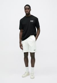 Férfi áll szemben, fekete EA7 logós pólót, fehér EA7 rövidnadrágot, fehér zoknit és fehér sportcipőt visel egy egyszerű háttér előtt.