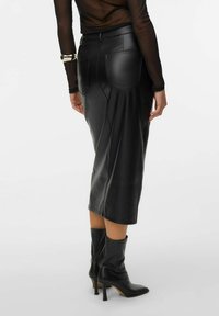 Zwarte leren midi-rok met gestructureerd ontwerp, zichtbare stiksels en twee achterzakken. G gedragen met zwarte hoge hakken.