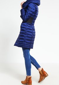 Navy pufferjack met capuchon en doorgestikte ontwerpen, gecombineerd met slim-fit jeans en bruine enkellaarsjes met een golvend patroon aan de zijkanten.