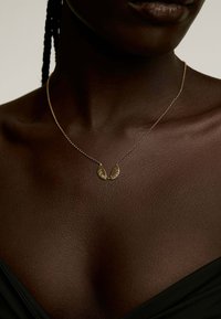 Collar de oro en forma de U con texto grabado, colgando de una delicada cadena, usado sobre piel oscura. Tela negra cae en el fondo.
