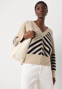 Beige neulepaita, jossa on tummansiniset vinoraitat; löysä istuvuus, neulottu rakenne, yhdistetty valkoisiin pö trousers ja vaaleankermanväriseen käsilaukkuun.