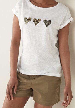 Persoon draagt een wit T-shirt met drie dierenprintharten en opgerolde mouwen, gecombineerd met beige shorts, staand tegen een lichte muur.