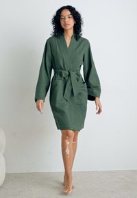 Robe verte texturée avec de longues manches et une ceinture à nouer, présentant une coupe jusqu'aux genoux et de subtiles rayures verticales. Design simple sans motifs.