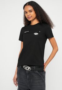 Čierne bavlnené tričko s krátkymi rukávmi, pásikovým detailom cez hruď a jemným grafickým dizajnom. Kombinované s tmavošedými džínsami a opaskom.