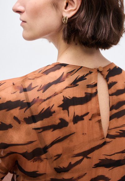 Γυναίκα με κοντά καστανά μαλλιά που φορά χρυσούς κρίκους σκουλαρίκια και καφέ μπλούζα με ρίγες τίγρης και λεπτομέρεια κουμπιών στην πλάτη.