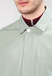 Mann trägt eine hellgrüne Jacke mit Kragen über einem weinroten Reißverschluss-Top mit schwarzen Streifen, zeigt Gesichtshaar und geschlossene Lippen.