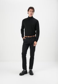 Czarny sweter z golfem, czarne spodnie, brązowy skórzany pasek i czarne buty wizytowe. Model stoi na neutralnym, białym tle.
