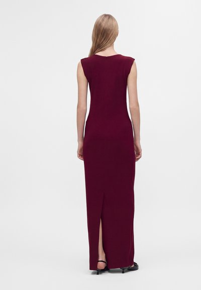 Femme aux longs cheveux blonds portant une robe longue sans manches rouge foncé avec une fente dans le dos et des talons noirs, debout devant un fond uni.
