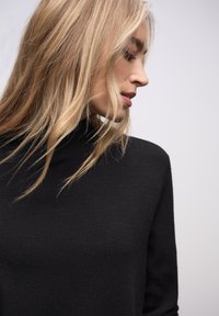 Pull à col roulé noir avec une texture douce et une coupe ajustée, présenté sur un fond neutre. De longs cheveux encadrent partiellement le col.