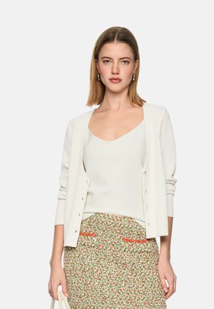 Woman wearing a white cardigan over a white tank top and a floral-patterned skirt, holding a white bag against a plain background.