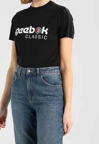Černé bavlněné tričko s bílým a červeným logem "Reebok CLASSIC", doplněné středně modrými džíny s klasickým střihem a pěti kapsami.