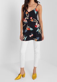 Czarna bluzka w kwiaty na cienkich ramiączkach z czerwonymi, różowymi i zielonymi wzorami. Połączona z białymi spodniami o dopasowanym kroju i żółtymi butami na słupku.