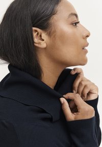 Mornarsko plava, rebrasta pletena turtleneck pulover sa preklopljenim ovratnikom, prikazano kako ga nositelj prilagođava oko vrata.