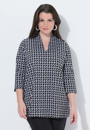 Frau mit langen dunklen Haaren, die große weiße Creolen und eine schwarz-weiße Bluse mit geometrischem Muster und Dreiviertelärmeln trägt.