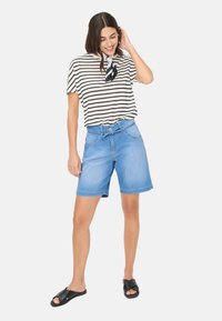 T-shirt à rayures à manches courtes beige et noir, associé à un short en denim bleu clair et des sandales noires à enfiler. Le mannequin pose avec une main dans les cheveux.