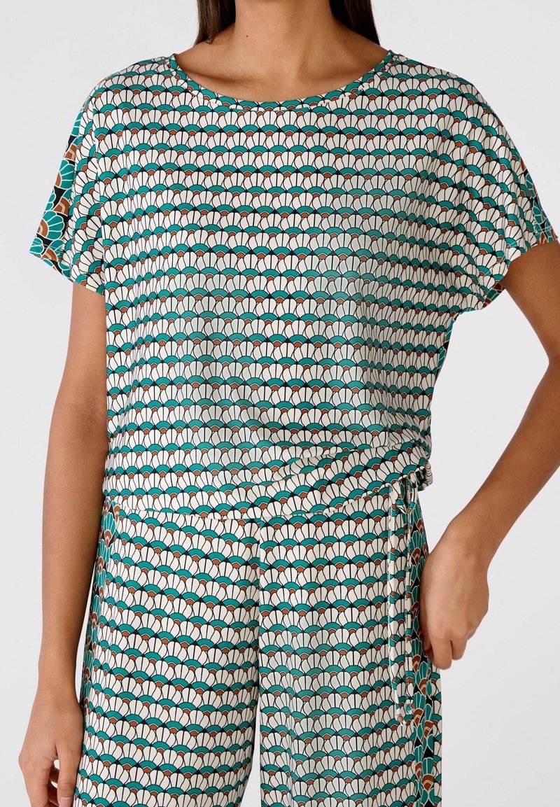 Femme portant un haut à motifs et un pantalon assorti avec des motifs en forme d'éventail bleu sarcelle, marron et blanc, debout avec une main sur la hanche.
