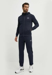 Felpa con zip colore navy e pantaloni abbinati realizzati in un tessuto morbido. Presenta un logo sul petto e sulla gamba sinistra dei pantaloni. Abbinato a scarpe da ginnastica bianche.