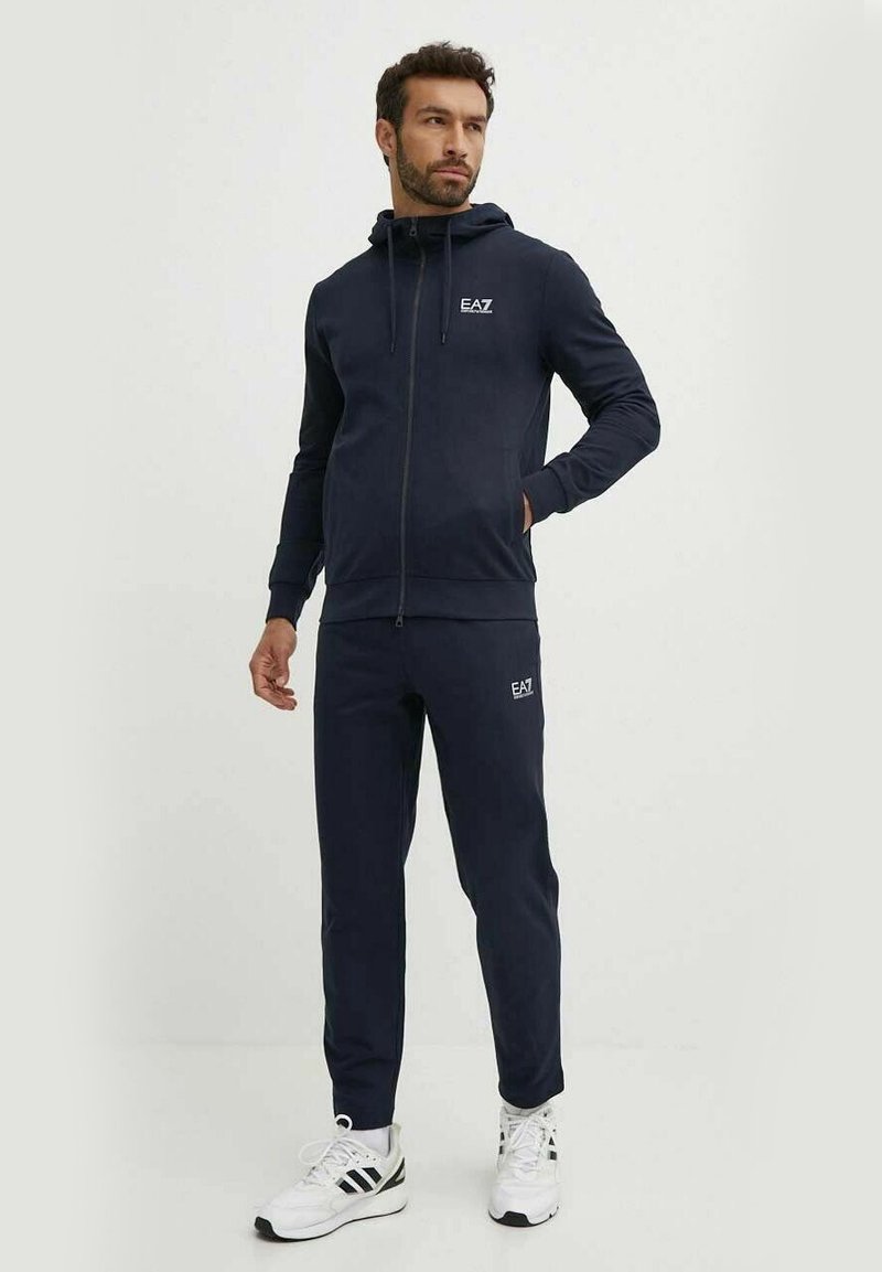 Felpa con zip colore navy e pantaloni abbinati realizzati in un tessuto morbido. Presenta un logo sul petto e sulla gamba sinistra dei pantaloni. Abbinato a scarpe da ginnastica bianche.