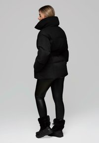 Černá péřová bunda s vysokým límcem, spárovaná s černými legínami z umělé kůže a černými kozačkami s kožešinovou podšívkou. Hladký materiál a polstrovaný design.